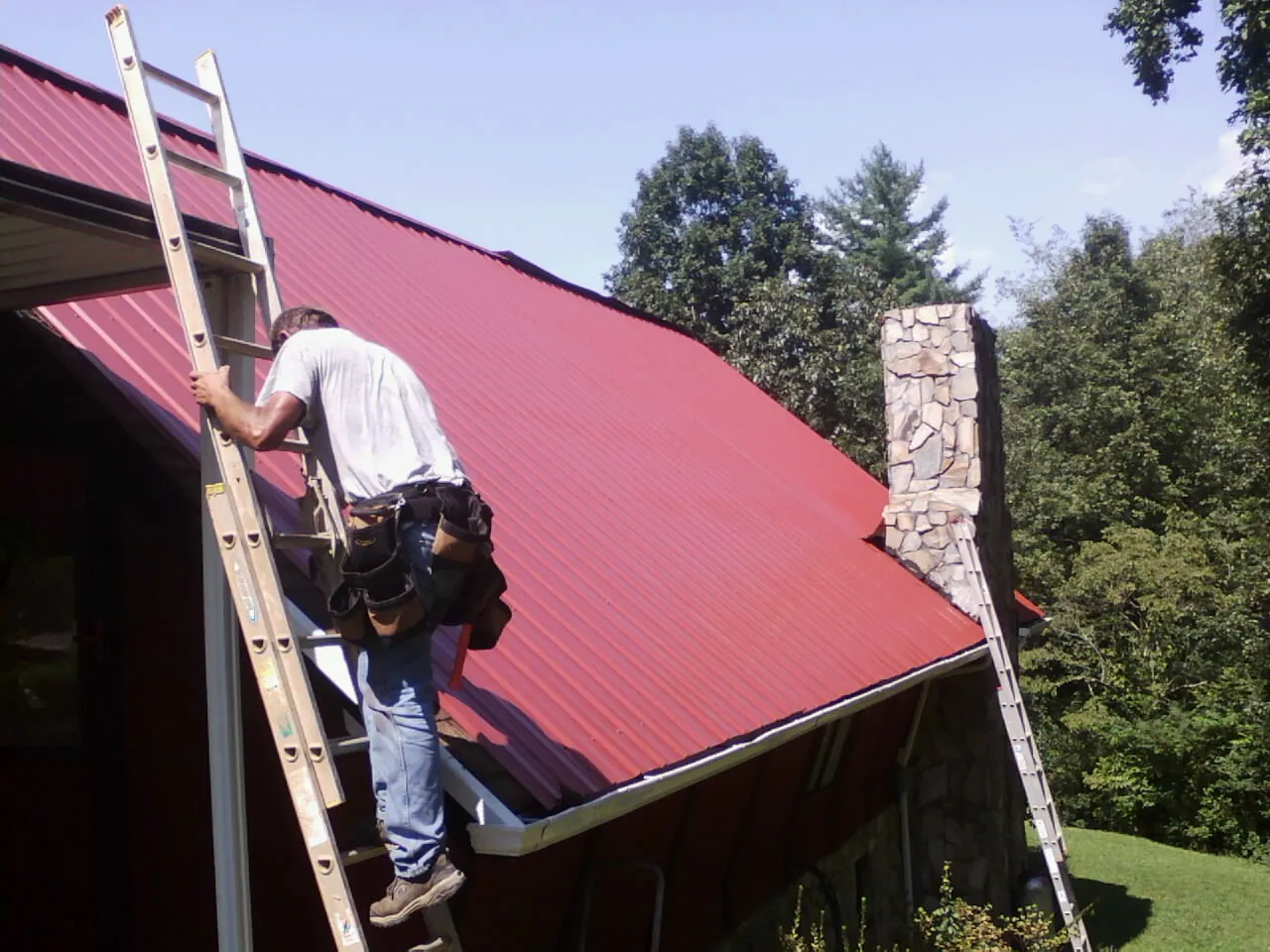 Roofer on a ladder beside a red metal roof and stone chimney — Soffit & Fascia Repair by Roofing Brothers in Bond